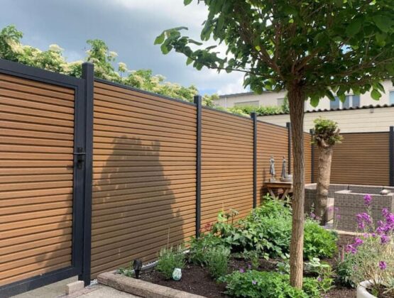 Strakke teak composiet schutting met zwarte aluminium palen in een verzorgde tuin met bloemen, boom en zitgedeelte.