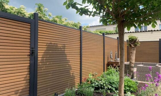 Strakke teak composiet schutting met zwarte aluminium palen in een verzorgde tuin met bloemen, boom en zitgedeelte.